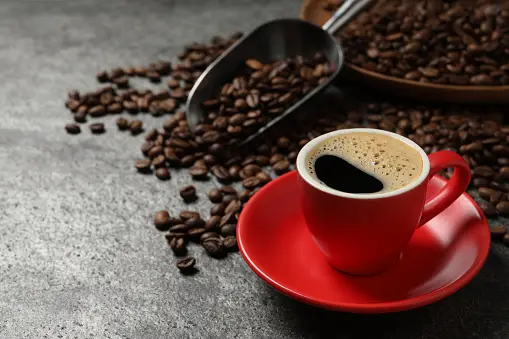 A jar of instant coffee powder with a steaming cup of coffee in the background.