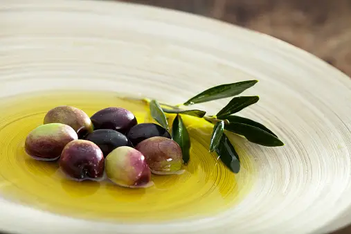 A glass bottle of Extra Virgin Olive Oil surrounded by fresh olives and herbs.
